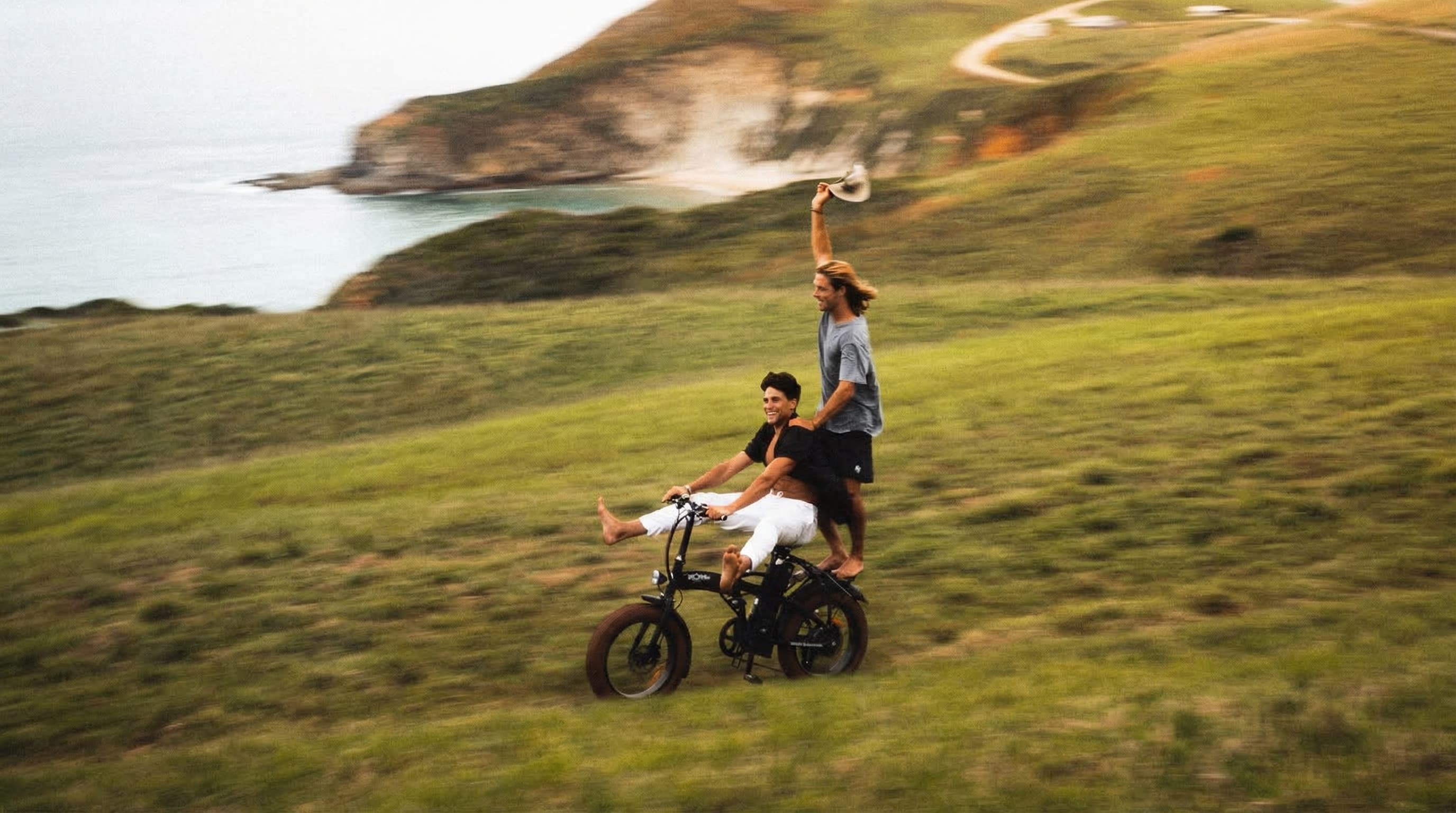 Two people riding an e-bike along a coastal hillside, joyful and free