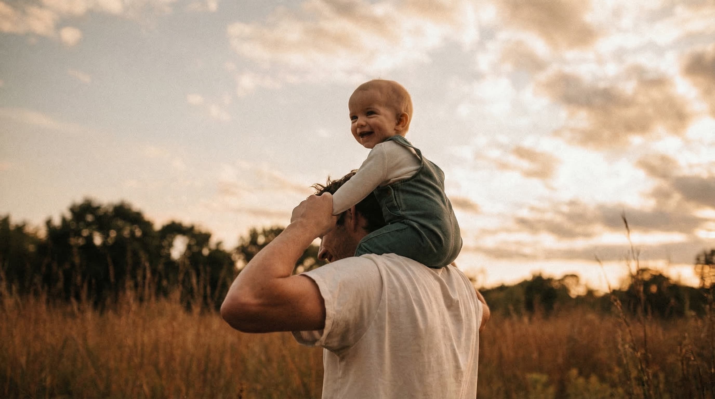 Man carrying a laughing baby on his shoulders in a golden field at sunset