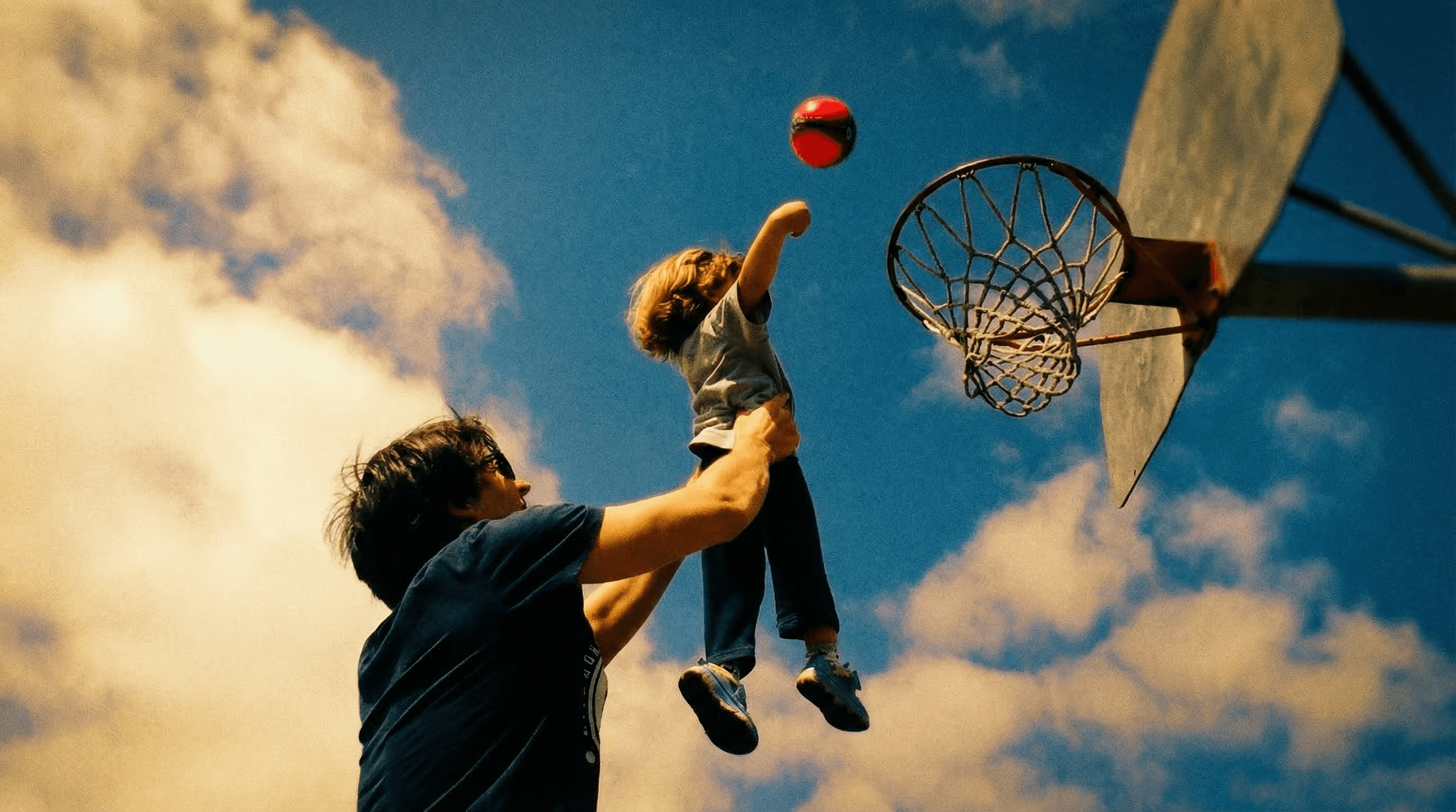 Father lifting child up to a basketball hoop against a dramatic sky